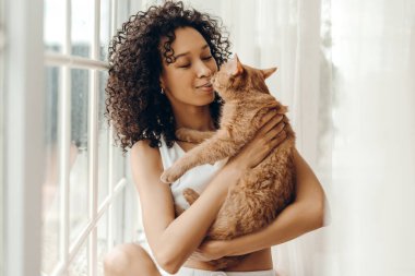 Sabahları gülümseyen kıvırcık saçlı Afro-Amerikan bir kadının kedi tutuş portresi. Evcil hayvanlar ve insanlar yaşam tarzı konsepti