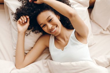 Kıvırcık saçlı, gülümseyen ve evde uyandıktan sonra yatakta gerinen neşeli Afro-Amerikan kadın manzarası. Yaşam tarzı kavramı