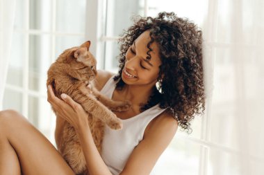 Memleketindeki büyük pencerenin yanında tüylü arkadaşıyla eğlenen mutlu Afrikalı Amerikalı genç bir kadının portresi. Evcil hayvanlar ve insanlar yaşam tarzı konsepti