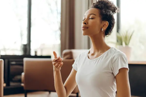 Genç Afrikalı Amerikalı kadın evde sessizce oturuyor, gözleri kapalı derin nefes alma alıştırması yapıyor. Sağlıklı, spor, meditasyon konsepti