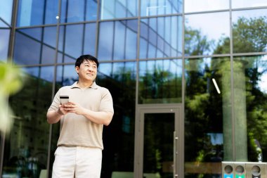Güneşli bir günde modern bir cam ofis binasının önünde dikilirken akıllı telefonu tutan gülümseyen bir işadamı.