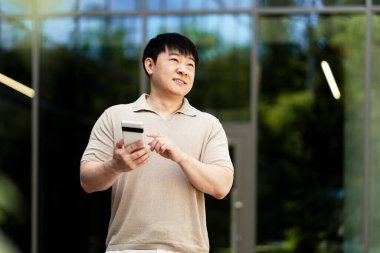 Gülümseyen işadamı akıllı telefon ve kredi kartıyla online ödeme yapıyor. Modern ofis binasının yanında duruyor.