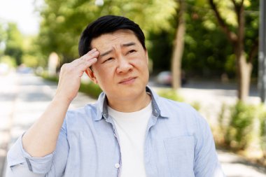 Baş ağrısından muzdarip Asyalı bir adam, eliyle alnına dokunuyor, ateşi ya da güneş çarpması geçiriyor, sağlıksız hissediyor, acı çekiyor, sokakta duruyor.