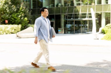 Modern ofis binasının dışında dizüstü bilgisayarla yürüyen kendine güvenen işadamı güneşli bir yaz gününde işe gidiyor.