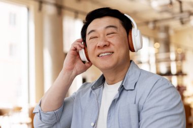 Gülümseyen Asyalı adam müzik dinlerken kablosuz kulaklık takıyor. Parlak, cezbedici bir kahve dükkanında. Mutluluk ve rahatlama yayıyor.