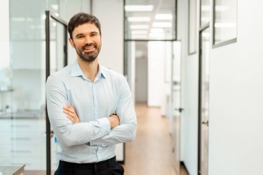 Kendine güvenen bir işadamının portresi kolları bağlı, modern bir ofis koridorunda duruyor ve dostça bir gülümseme, profesyonelliği ve liderliği somutlaştırıyor. Reklam kavramı