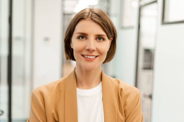 Modern bir ofis koridorunda özgüvenle gülümseyen başarılı bir iş kadınının portresi şık bir spor ceket ve beyaz tişört giyerek profesyonelliği yansıtıyor. Başarılı iş konsepti