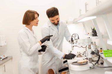 Laboratuvar önlüğü ve siyah eldiven giyen iki diş teknisyeni modern bir değirmen makinesi üzerinde çalışıyorlar. Laboratuvar ortamındaki ekipmanlara işaret ediyorlar. Tıbbi konsept