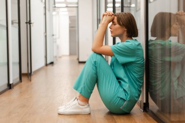 Turkuaz önlüklü üzgün kadın doktor bir hastane koridorunda yerde oturuyor, ara veriyor ve kafasını elinde tutuyor, yorgunluk ve stres yaşıyor. Kötü haber konsepti