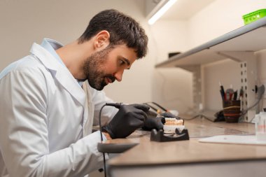 Siyah eldiven ve laboratuvar önlüğü giyen konsantre bir diş teknisyeni diş macunu kalıbı üzerinde dikkatle çalışmak için özel bir alet kullanıyor.