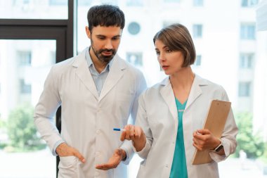 İki doktor teşhisi tartışıyor, biri elinde bir not panosu, diğeri ise kalem, işbirliği ve hastane ortamında tıbbi uzmanlık ile el kol hareketi yapıyor. Tartışma konsepti