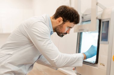 Laboratuvar önlüğü ve eldiven giyen bilim adamı, muhtemelen modern bir laboratuvar ortamında deney ya da analiz yapan karmaşık ekipmanları dikkatlice kullanıyor. Bilim konsepti