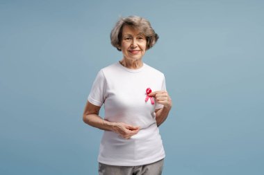 Beyaz tişörtüne pembe kurdele takmış gülümseyen son sınıf öğrencisi kadın göğüs kanseri farkındalığını destekliyor açık mavi arka planda poz veriyor.