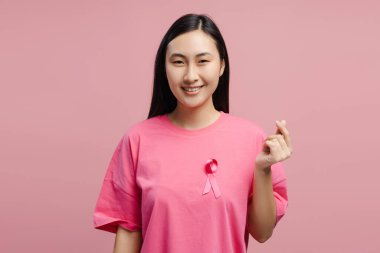 Pembe kurdeleli pembe tişört giyen gülümseyen Asyalı kadın göğüs kanseri farkındalığını destekleyen parmaklarıyla küçük kalp işaretleri gösteriyor, pembe stüdyo arka planında izole edilmiş poz veriyor. Sağlık hizmeti kavramı