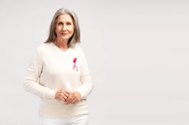 Beyaz kazaklı, pembe kurdeleli, gülümseyen bir kadın beyaz arka planda kameraya bakıyor. Göğüs kanseri kavramı