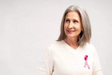 Gülümseyen, mutlu, olgun, beyaz saçlı, pembe kurdeleli sıradan beyaz elbiseli bir kadının portresi. Fotokopi alanına bakıyor, beyaz arka planda tek başına duruyor. Sağlık hizmeti kavramı