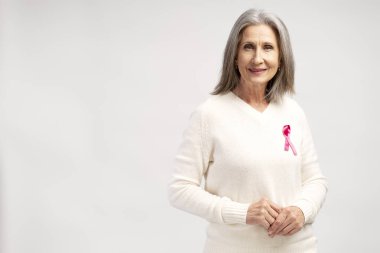 Gülümseme portresi, beyaz günlük kıyafetler ve pembe kurdele takan, beyaz arka planda izole edilmiş poz veren, yaşlı, pozitif gri saçlı kadın. Göğüs kanseri farkındalığı konsepti
