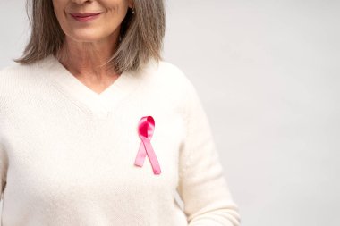 Yakın plan, beyaz kazak giyen, pembe kurdeleli, seçici odaklı, beyaz arka planda yalnız poz veren yaşlı gri saçlı kadın portresi. Göğüs kanseri farkındalığı konsepti