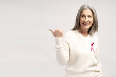 Gülümseyen, yaşlı, gri saçlı, pembe kurdeleli beyaz kazak giyen, fotokopi alanını işaret eden, beyaz arka planda izole edilmiş poz veren bir kadın portresi. Reklam, göğüs kanseri farkındalığı konsepti
