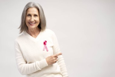 Gülümseyen, neşeli, yaşlı, beyaz saçlı, pembe kurdeleli beyaz kazak giyen, fotokopi alanını işaret eden beyaz arka plandaki kameraya bakan kadının portresi. Göğüs kanseri kavramı