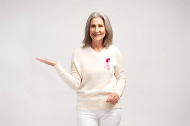 Kendinden emin, mutlu, gülümseyen gri saçlı, pembe kurdeleli beyaz kazak giyen, fotokopi alanında ellerini gösteren, kameraya bakan, beyaz arka planda izole edilmiş poz veren bir kadın. Brest kanseri, destek kavramı