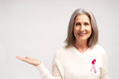 Yakından, mutlu, gülümseyen gri saçlı, pembe kurdeleli beyaz kazak giyen, fotokopi alanında ellerini gösteren, kameraya bakan, beyaz arka planda izole edilmiş poz veren bir kadın. Brest kanseri, destek kavramı