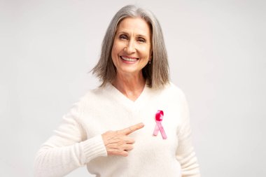 Beyaz kazak giyen, pembe kurdeleli, gülümseyen, gri saçlı bir kadının portresi beyaz arka planda yalnız poz veren kameraya bakıyor. Göğüs kanseri farkındalığı konsepti