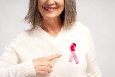 Yakın plan, beyaz kazak giyen gülümseyen gri saçlı kadının portresi, pembe kurdeleyi işaret eden, seçici odaklı, beyaz arka planda izole edilmiş. Sağlık hizmetleri, göğüs kanseri kavramı