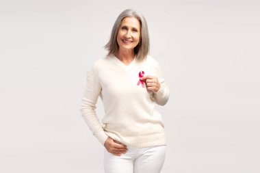 Gülümseyen, beyaz günlük kıyafetler giyen, elinde pembe kurdele tutan, kameraya bakan, beyaz arka planda izole bir şekilde poz veren çekici bir kadın portresi. Göğüs kanseri dövüş konsepti