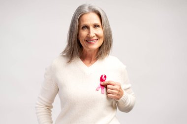 Gülümseyen, güzel, mutlu, son sınıf öğrencisi beyaz günlük kıyafetler giyen, pembe kurdeleli, kameraya bakan, beyaz arka planda izole edilmiş poz veren bir kadının portresi. Göğüs kanseri dövüş konsepti