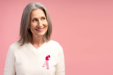Gülümseyen, mutlu, pozitif olgun bir kadının portresi. Pembe kurdeleli şık beyaz bir kazak giyen, başka tarafa bakan, pembe arka planda izole edilmiş, fotokopi çeken bir kadın. Göğüs kanseri kavramı