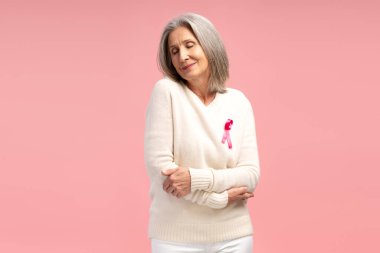 Güzel, gülümseyen olgun bir kadının portresi pembe kurdeleli beyaz bir kazak giyen, pembe arka planda izole bir şekilde kendine sarılan. Destek, göğüs kanseri konsepti