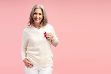 Mutlu, güzel, gri saçlı, pembe kurdeleli beyaz kazak giyen, pembe arka planda izole edilmiş kameraya bakan, fotokopi çeken bir kadın. Reklam, göğüs kanseri konsepti