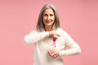 Güzel, gülümseyen gri saçlı bir kadının portresi pembe kurdeleli sıradan bir süveter giyiyor, kameraya bakıyor, pembe arka planda izole bir şekilde poz veriyor. Göğüs kanseri kavramı