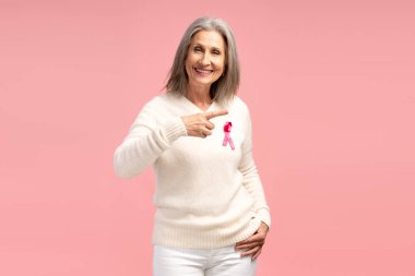 Mutlu yaşlı, gri saçlı, beyaz kazaklı, pembe kurdeleli bir kadının portresi fotokopi alanını işaret ediyor, pembe arka planda izole edilmiş poz veren kameraya bakıyor. Göğüs kanseri farkındalığı konsepti