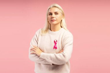 Kollarını çapraz bağlamış pembe kurdele takan, meme kanseri farkındalığını destekleyen kendinden emin olgun bir kadının stüdyo portresi.