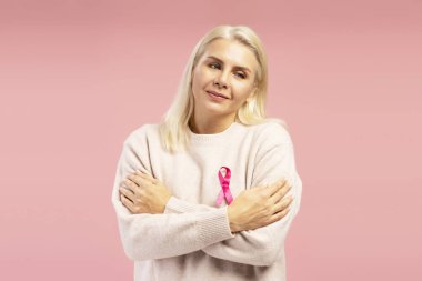 Göğüs kanserini destekleyen kolları çapraz ve pembe kurdeleli gülümseyen olgun bir kadının stüdyo portresi.