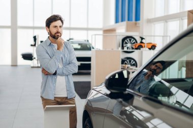 Düşünceli erkek müşteri yeni bir araba seçiyor, düşünceli bir şekilde çenesine dokunuyor ve araçlarla dolu modern araba galerisinde dikiliyor.