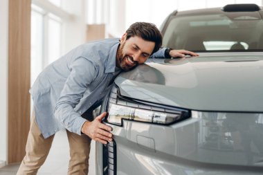 Mutlu, olgun bir erkek müşteri yeni elektrikli arabasını galeride kucaklıyor. Satışından duyduğu heyecanı ve memnuniyeti dile getiriyor. Otomatik alışveriş kavramı