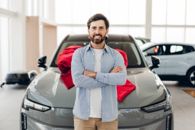 Mutlu müşteri, yeni gri arabasının önünde kırmızı bir fiyonk takmış bir araba galerisinin önünde gururla ve neşeyle alışverişini kutlarken çapraz kollarla gülümsüyor.