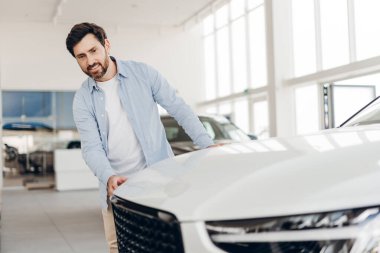 Otomobil galerisinde beyaz bir arabayı inceleyen, kaputa dokunan ve diğer araçlarla çevrili olan alımını düşünen mutlu bir adam. Ulaşım kavramı