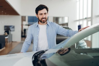 Mavi gömlekli sakallı adam araba galerisindeki yeni arabayı inceliyor, aracın kaputuna dokunuyor ve gülümsüyor, satın alma konusunda karar veriyor, satış temsilcisi ona yardım ediyor. Ulaşım kavramı