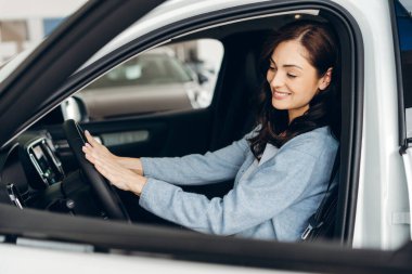 Mutlu bayan müşteri araba galerisinde yeni arabasında oturuyor, direksiyonu tutuyor ve gülümsüyor, satın almadan önce aracı test ediyor. Ulaşım kavramı