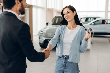 Gülümseyen kadın araba satıcısıyla el sıkışıyor ve galeride durup başarılı araba alımını kutlarken yeni araba anahtarları alıyor. Ulaşım kavramı