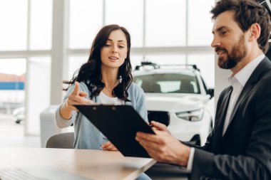 Profesyonel pazarlamacı, modern otomobil galerisinde masa başında oturmuş yeni otomobil alımını tartışırken kadın müşteriye belgeleri işaret ederek sözleşmenin detaylarını gösteriyor.