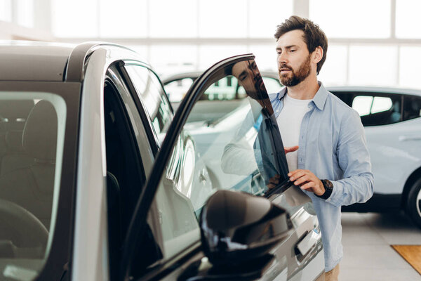 Bearded man opening car door, examining interior features and assessing condition of vehicle in auto showroom, thoughtfully considering decision to purchase new automobile
