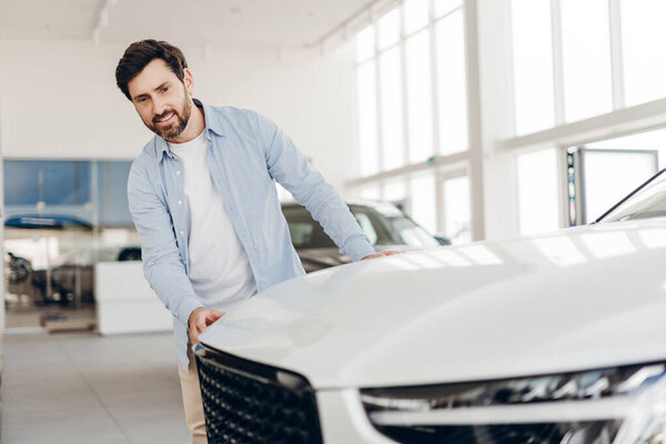 Счастливчик осматривает белый автомобиль в автосалоне, трогает капот и рассматривает его покупку, окруженный другими автомобилями. Транспортная концепция