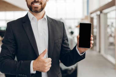 Gülümseyen araba satıcısı cep telefonunu boş ekranla gösteriyor ve başparmağını kaldırıyor, otomobil almak veya kiralamak için yeni mobil uygulamaları öneriyor