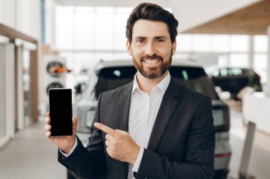 Gülümseyen araba satıcısı bir araba galerisinde dururken boş ekranla akıllı telefonu işaret ederek araçları satın almak için uygun internet hizmetleri sunuyor. Çevrimiçi teknoloji kavramı