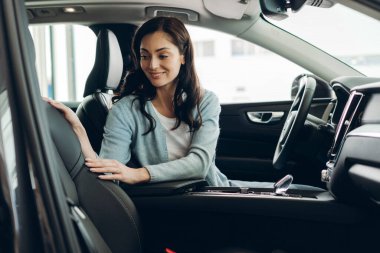 Gülümseyen kadın deri koltuğa dokunan yeni otomobilin içini inceliyor araba galerisinde araç seçiyor satın alma sürecinin tadını çıkarıyor satın alma konusunda karar veriyor.
